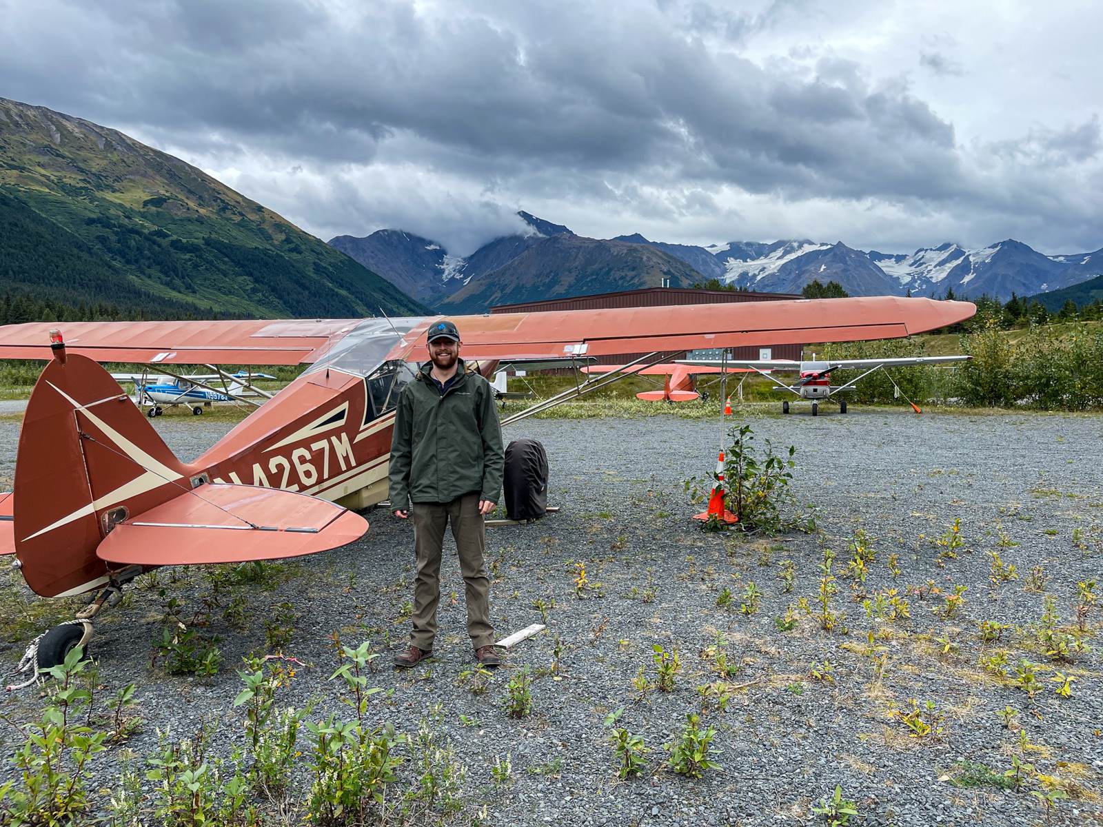 Standing beside a red bush plane on a gravel airstrip, snow-streaked mountains and low clouds behind