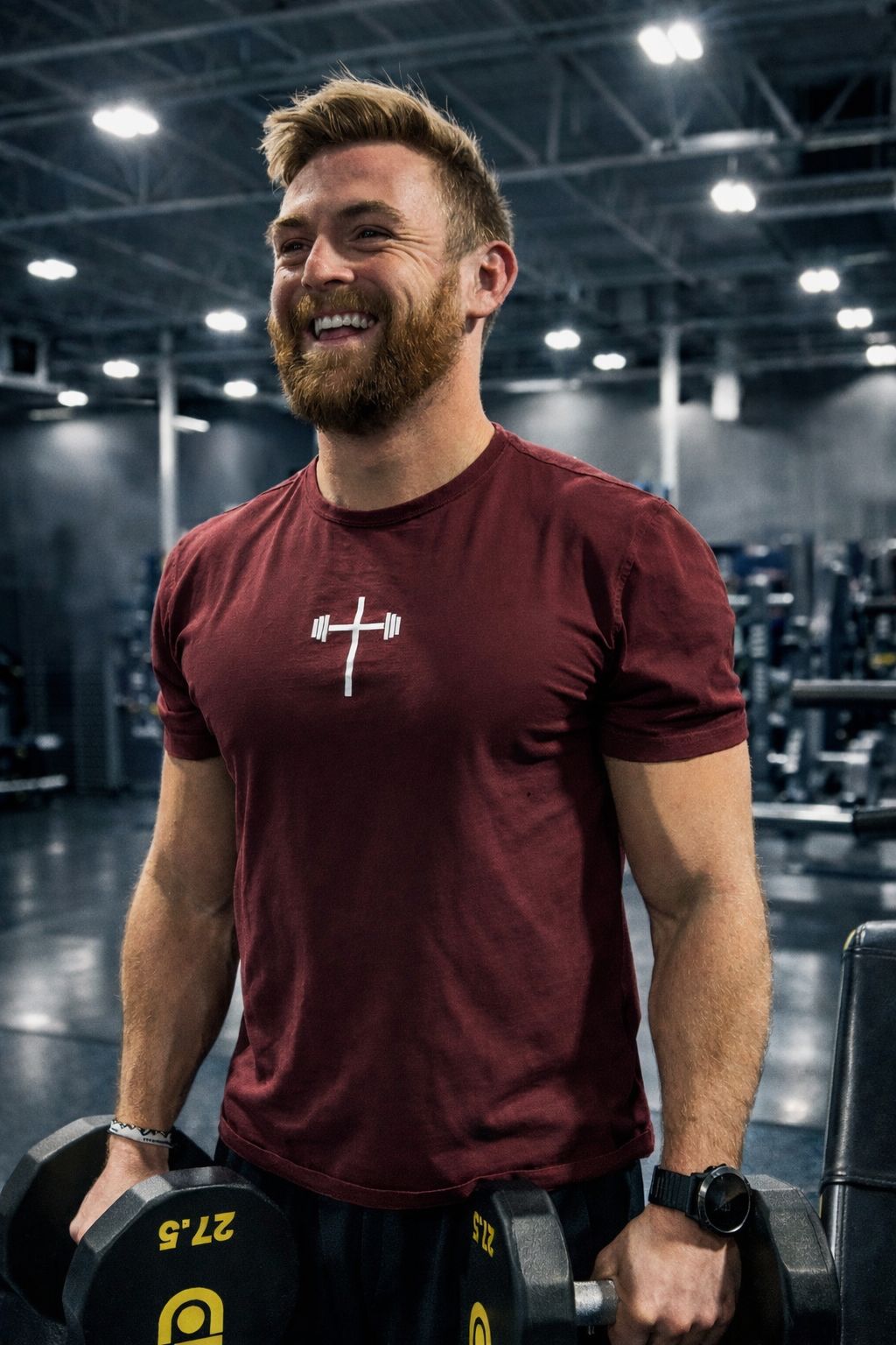 Will, smiling mid-set with a pair of dumbbells in the gym, wearing a cross-and-barbell tee