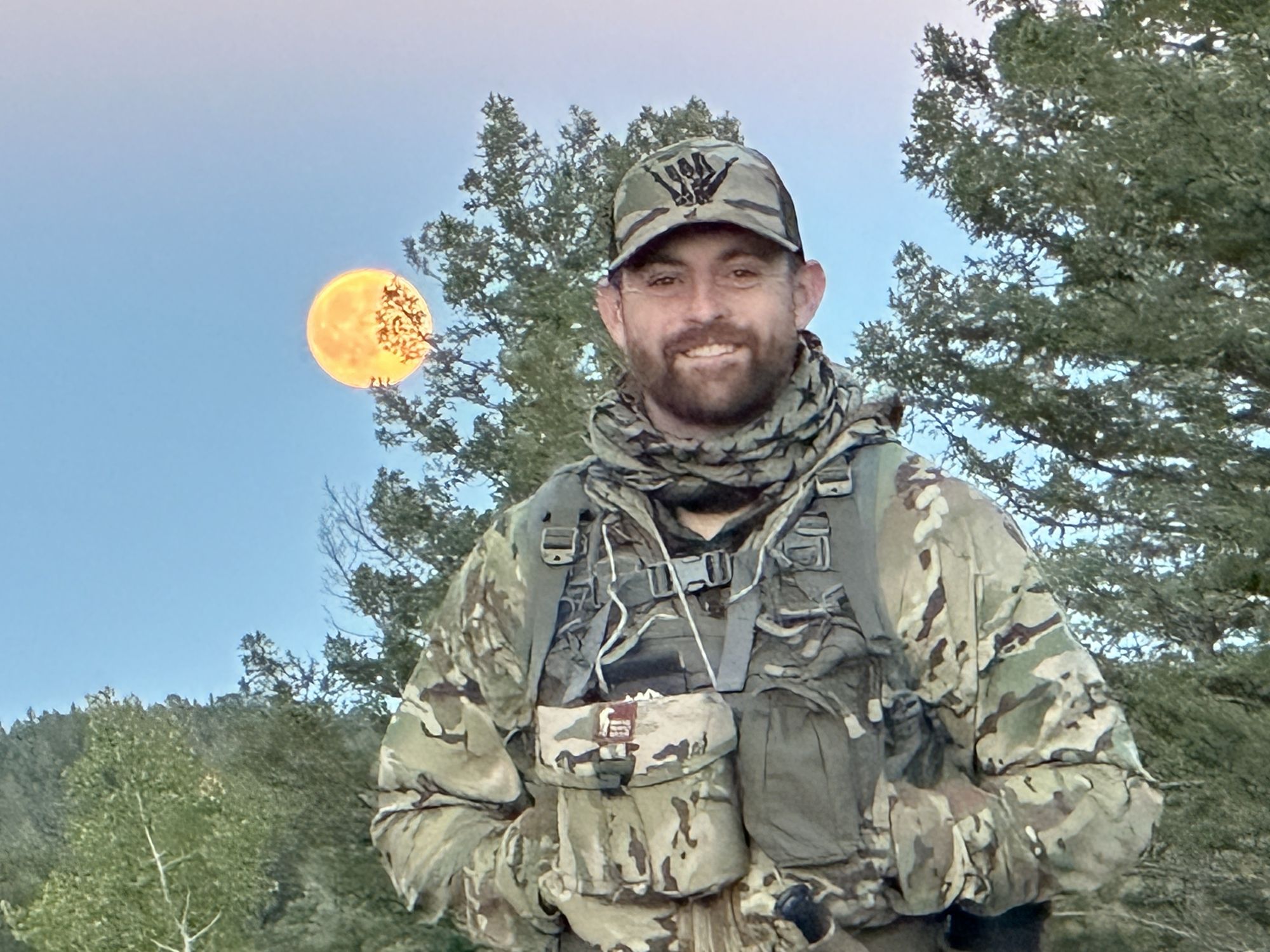 Will on a ruck in the mountains at dusk, smiling, with a full moon rising through the pines behind him