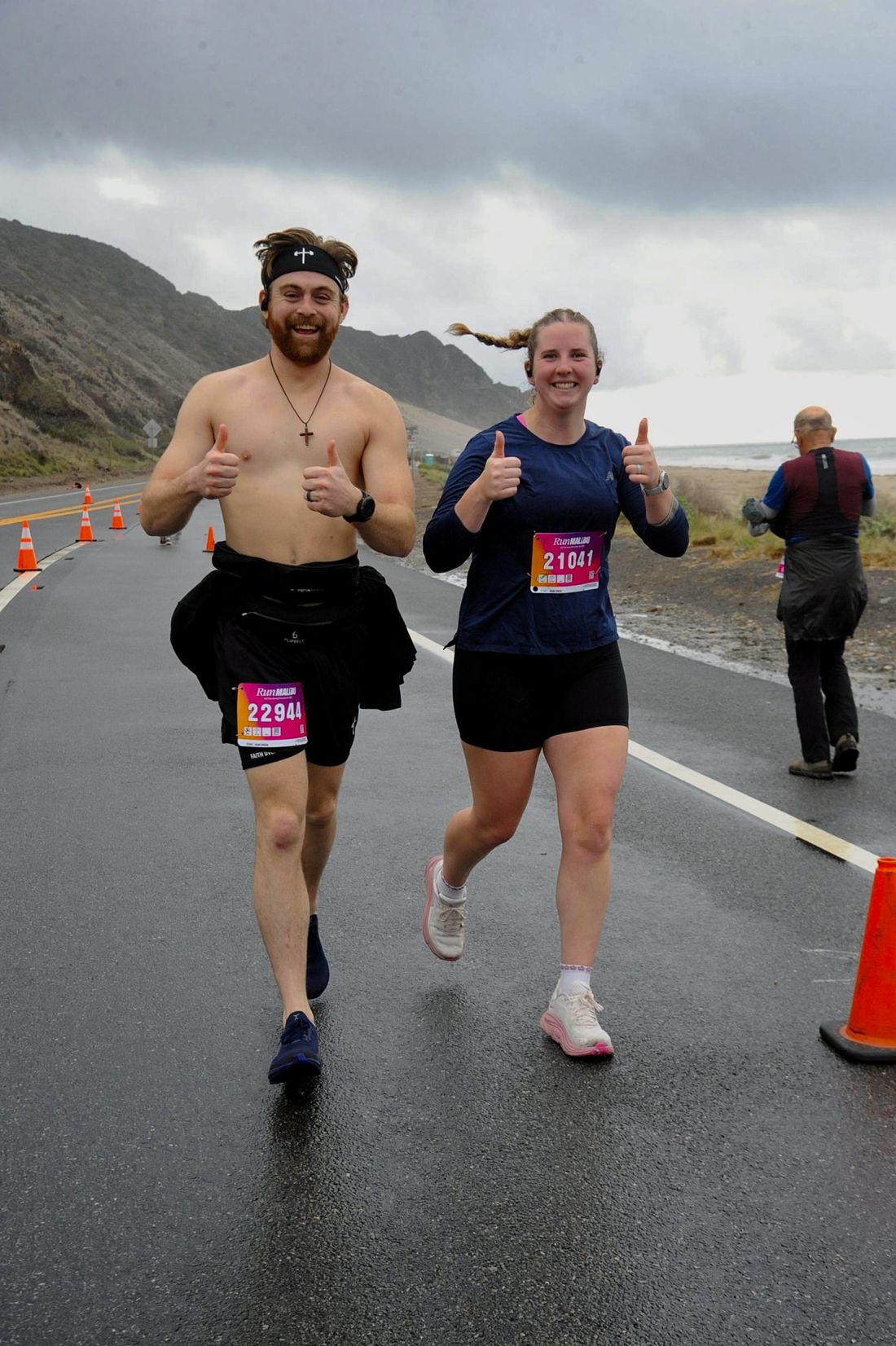 A husband and wife running a rainy half marathon along the Malibu coast, both grinning with thumbs up